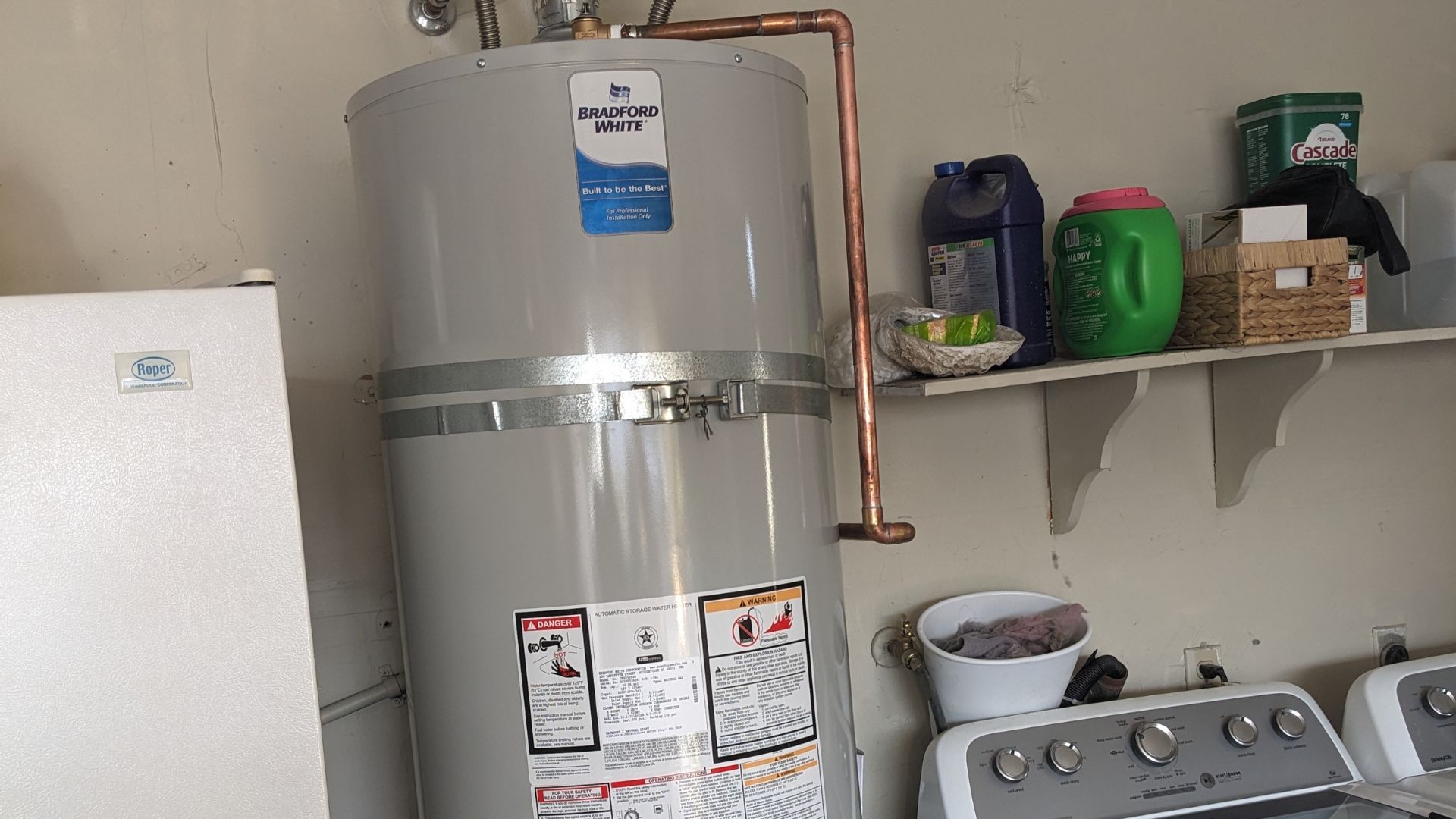 Water heater in a laundry room with copper pipes, a washing machine, and shelves with cleaning supplies.