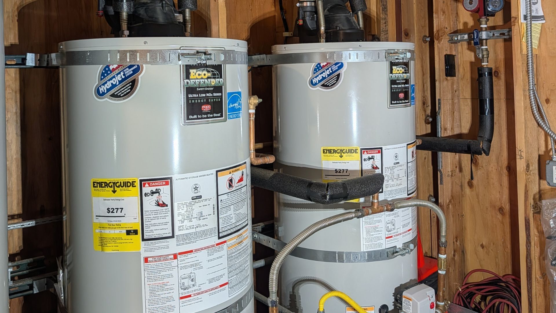 Two silver water heaters in a utility room. Pipes and wiring are visible, mounted against wood paneling.