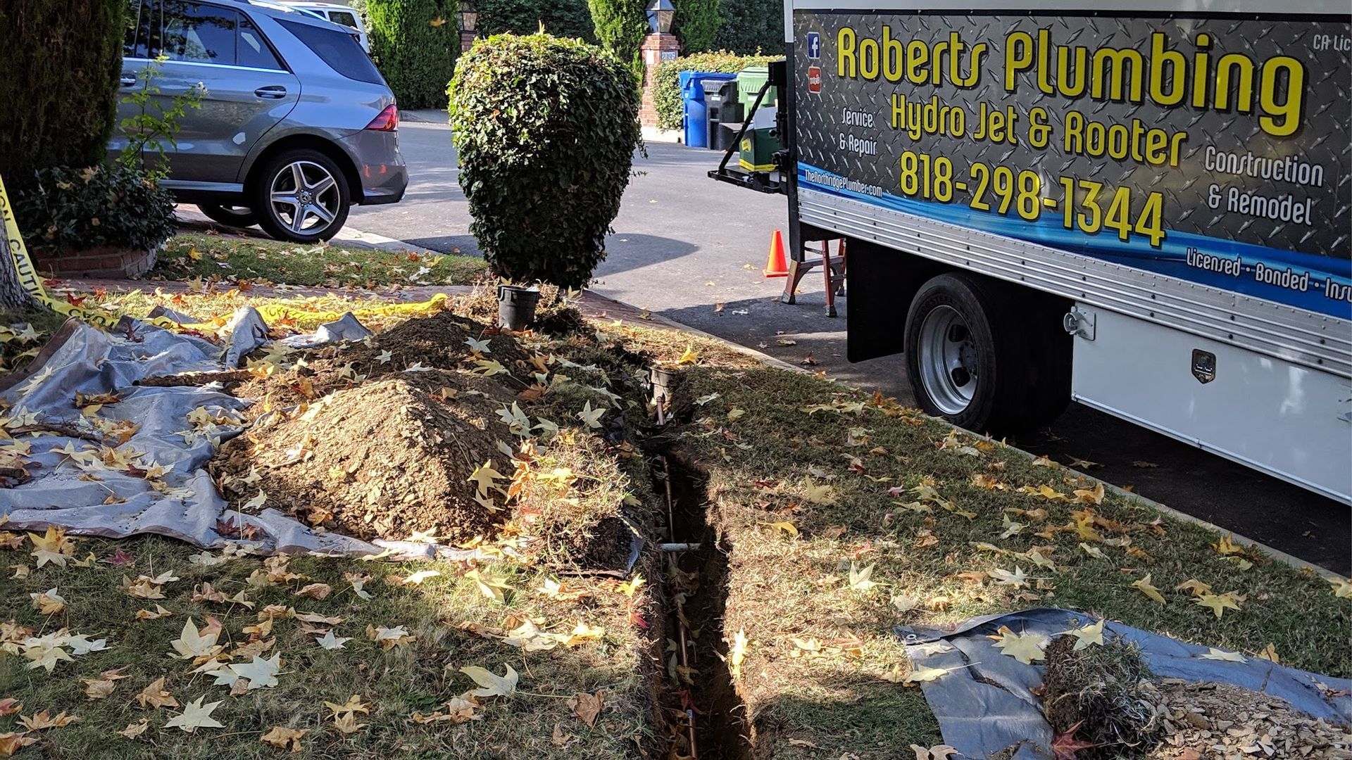 Trench dug in a lawn next to a Roberts Plumbing truck. A car and a small bush are in the background.