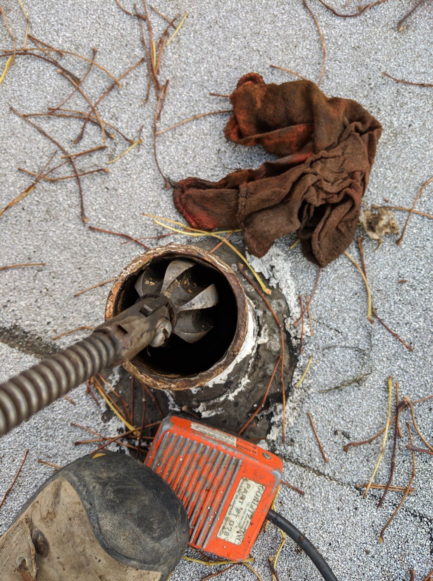 A drain being cleared with a mechanical snake on a roof. A brown rag and tool box are nearby.