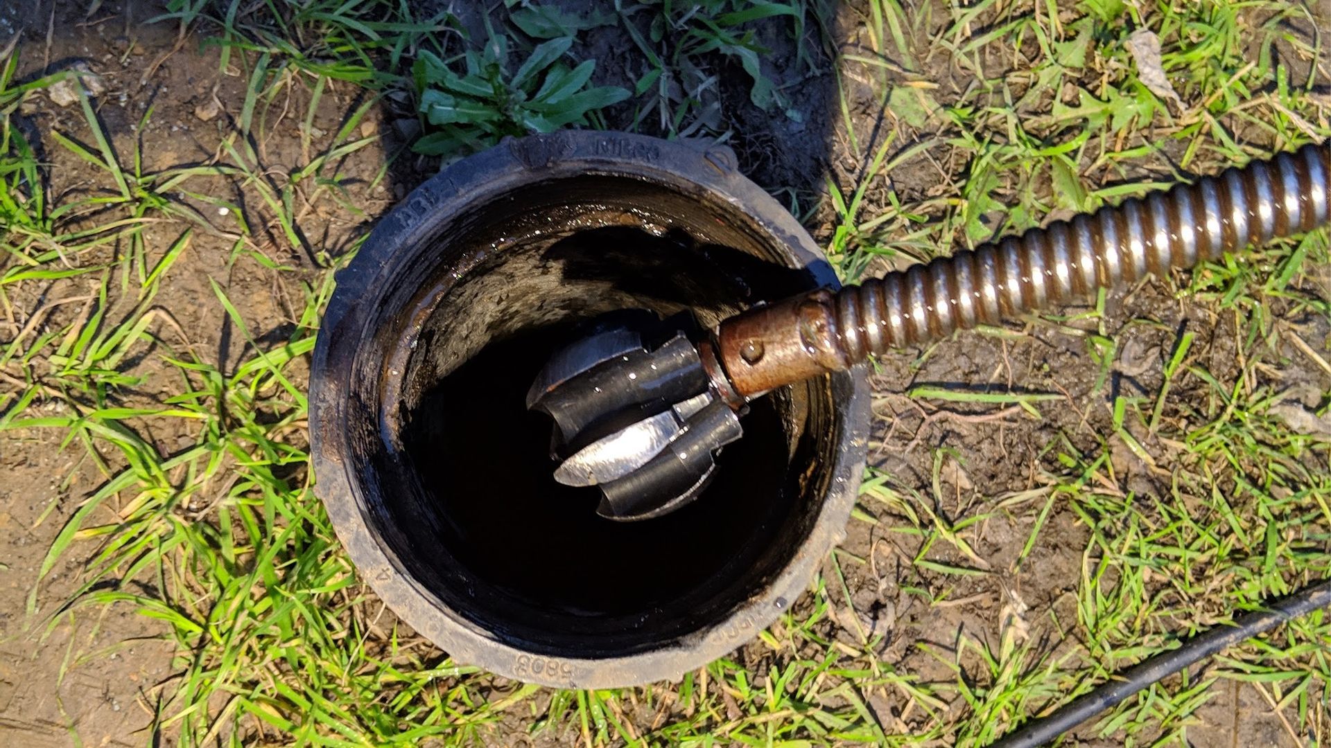Drain cleaning tool inside a dark pipe, surrounded by grass and dirt.