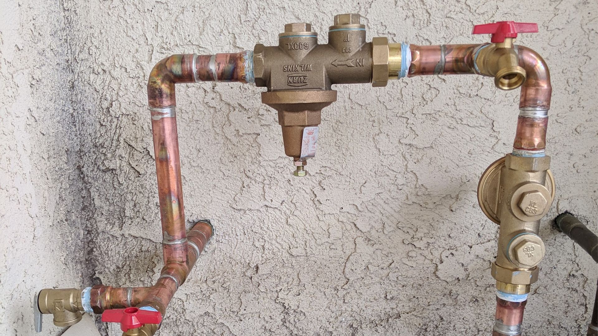 Copper pipes with valves and a pressure regulator against a textured wall.