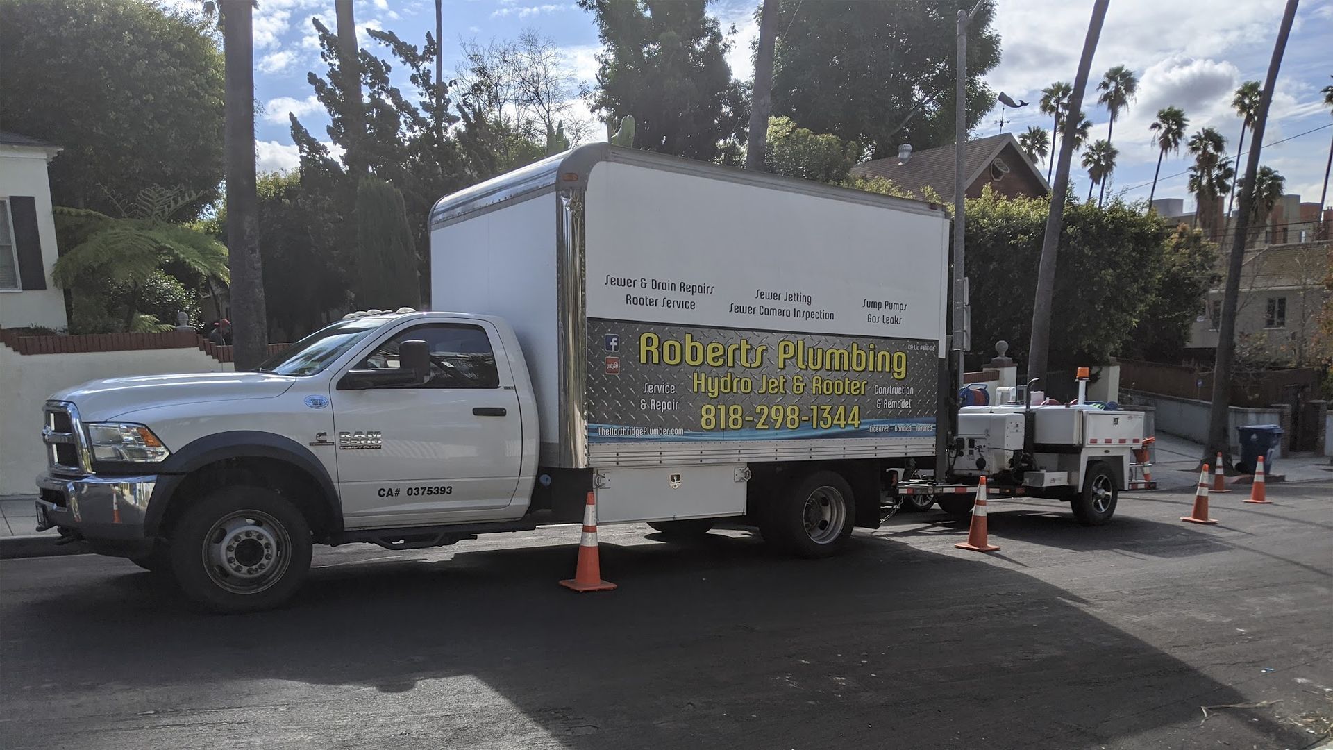 White truck with box trailer parked on the street with traffic cones; Robert's Upholstery sign. Trees in the background.