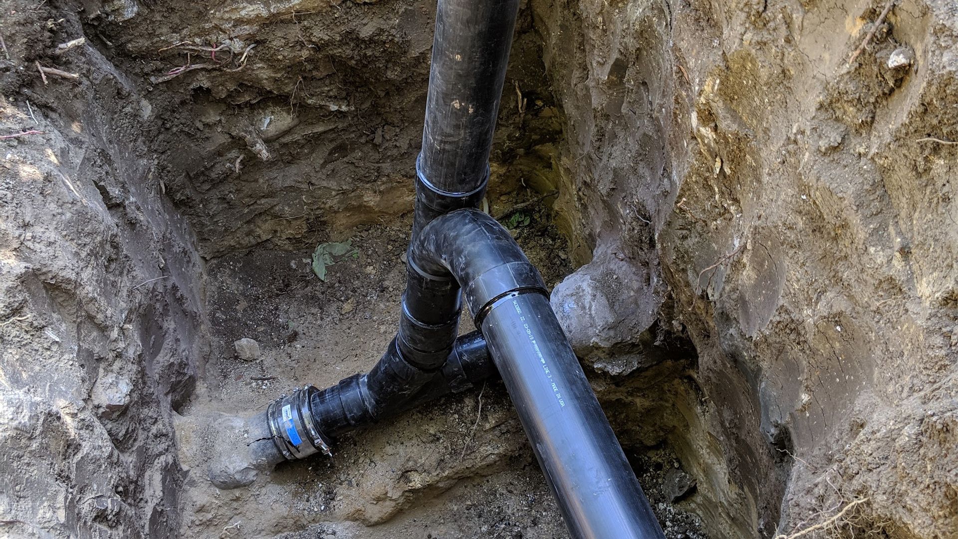 Black plumbing pipes in a dirt trench, branching in a Y-shape; construction.