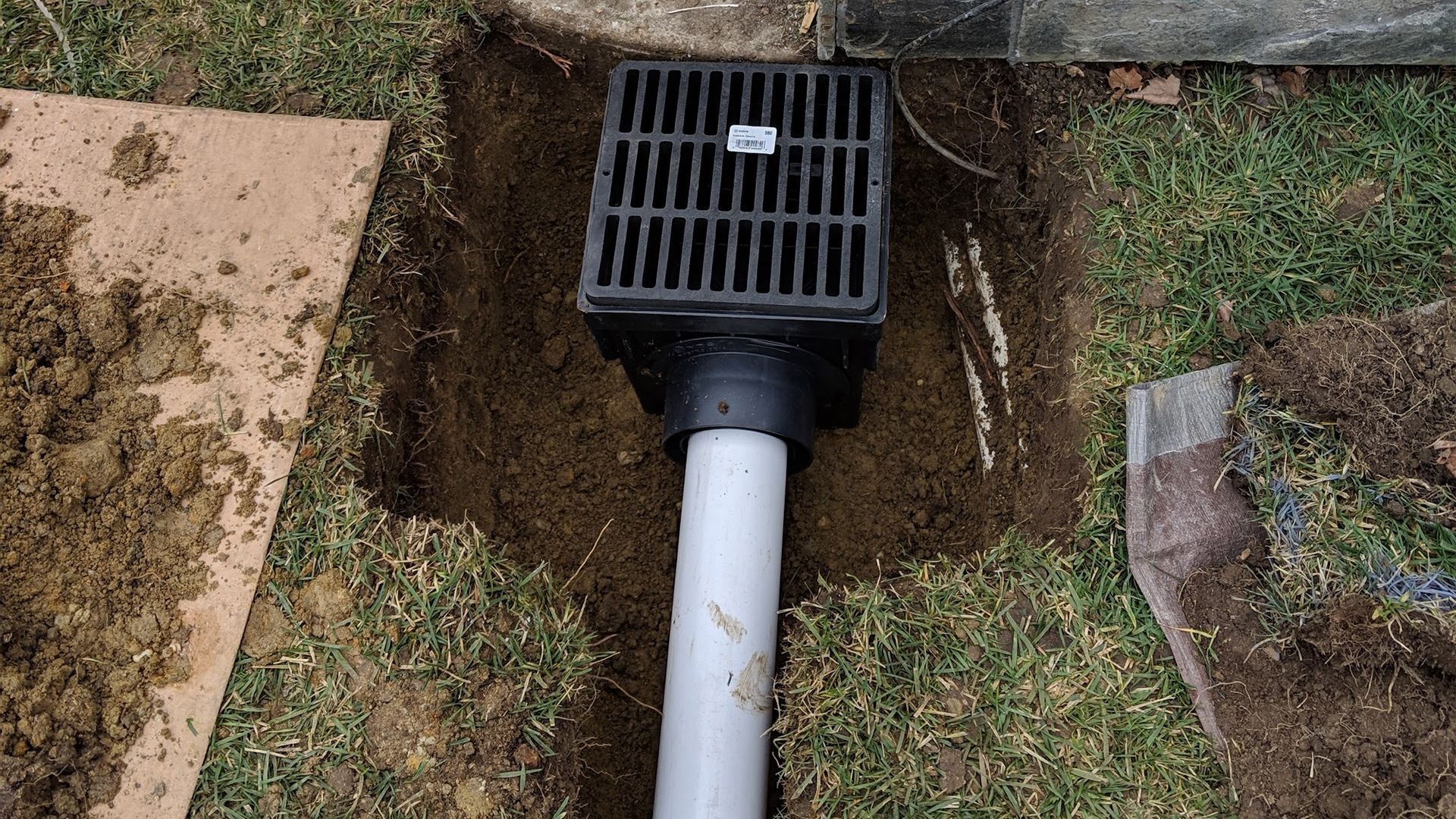 Drainage system installation in a lawn; black grate, white pipe in a trench.