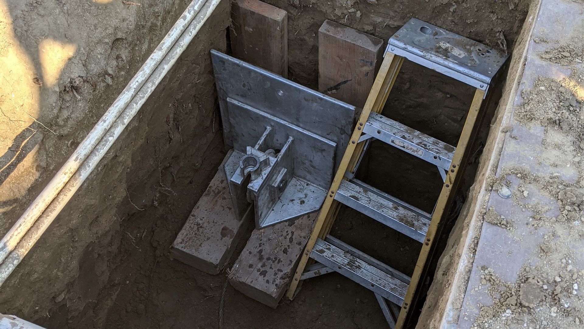 Ladder and metal structure inside a rectangular excavation pit, with wooden supports.