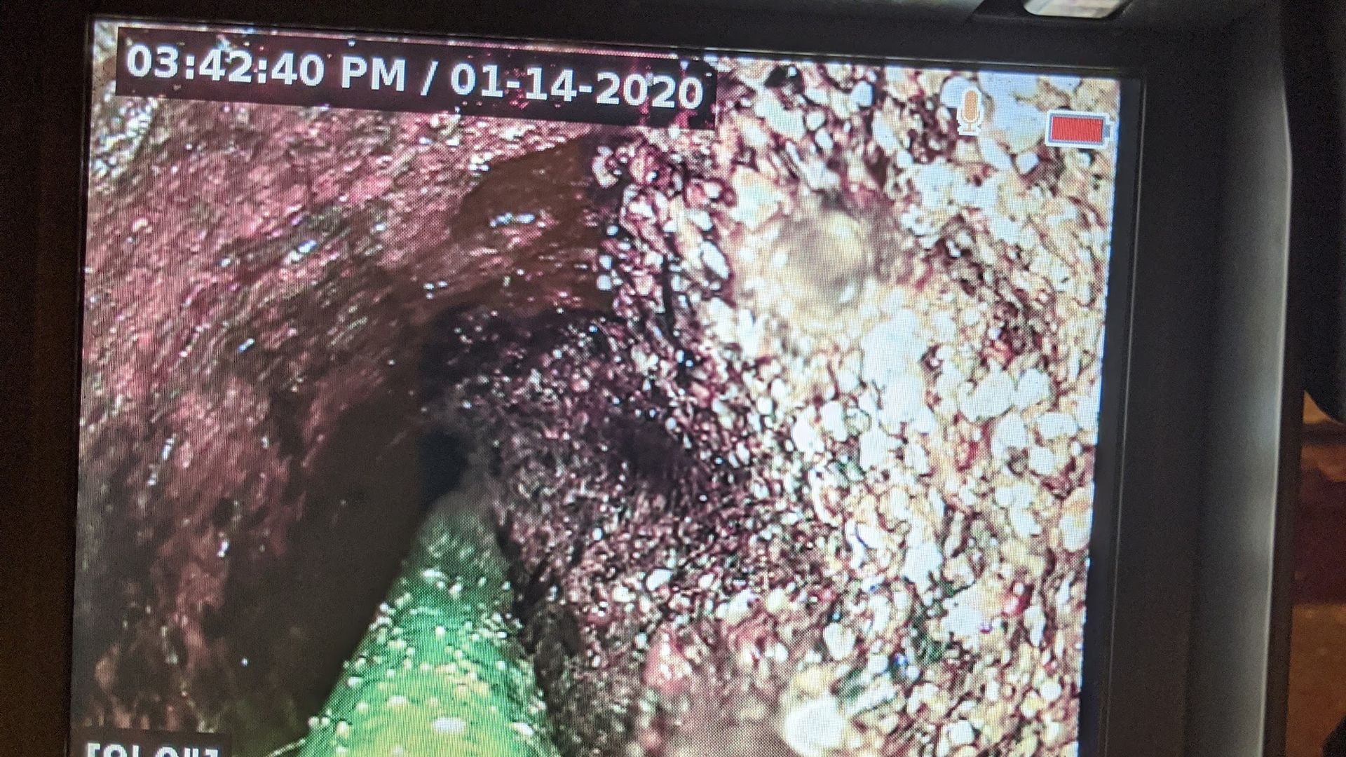 View inside a pipe, likely a drain, showing dark, wet surfaces and small stones with a green object in the bottom left.