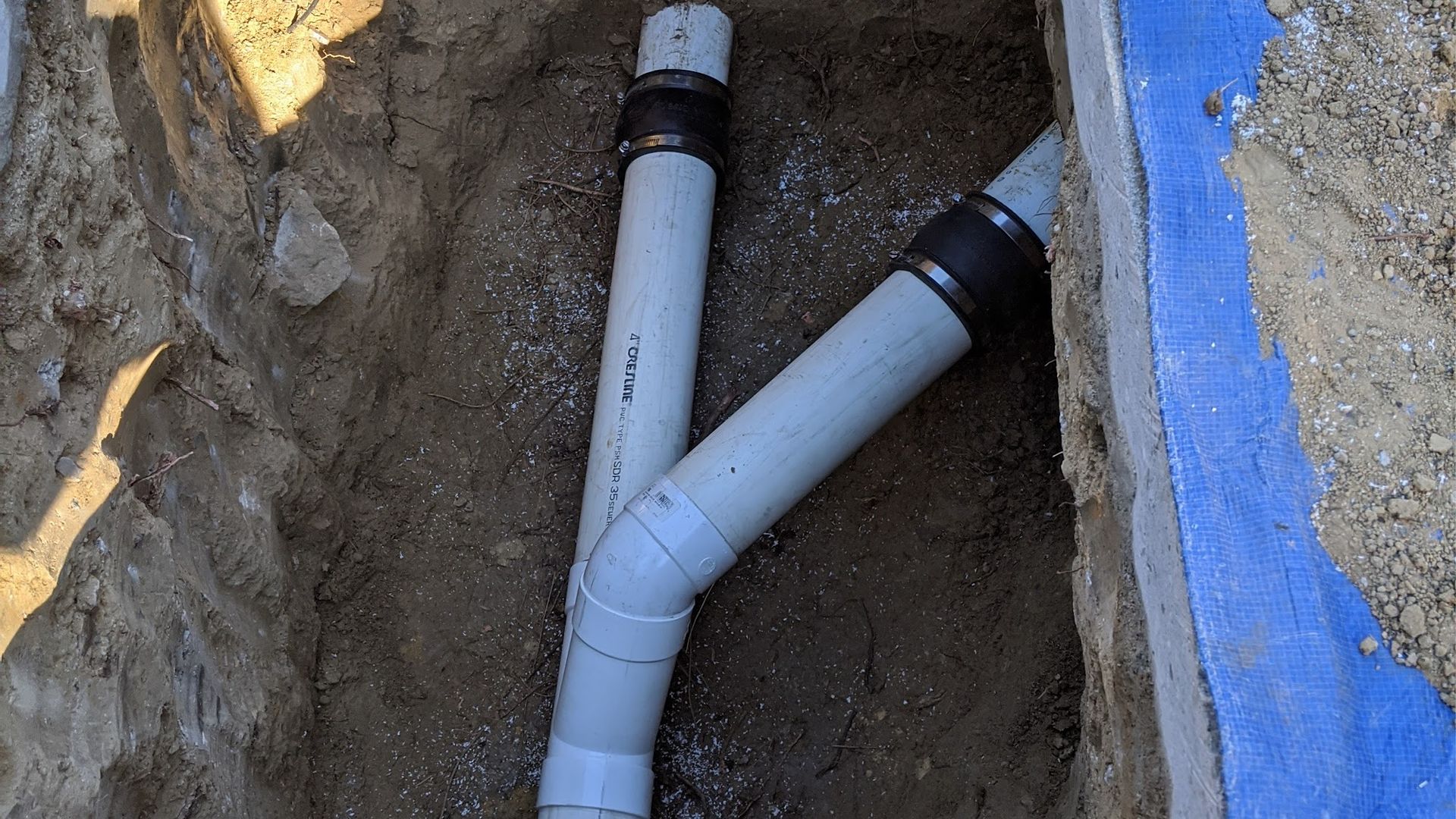 Two gray PVC pipes joined by a Y-fitting in a trench. Blue concrete edge visible.