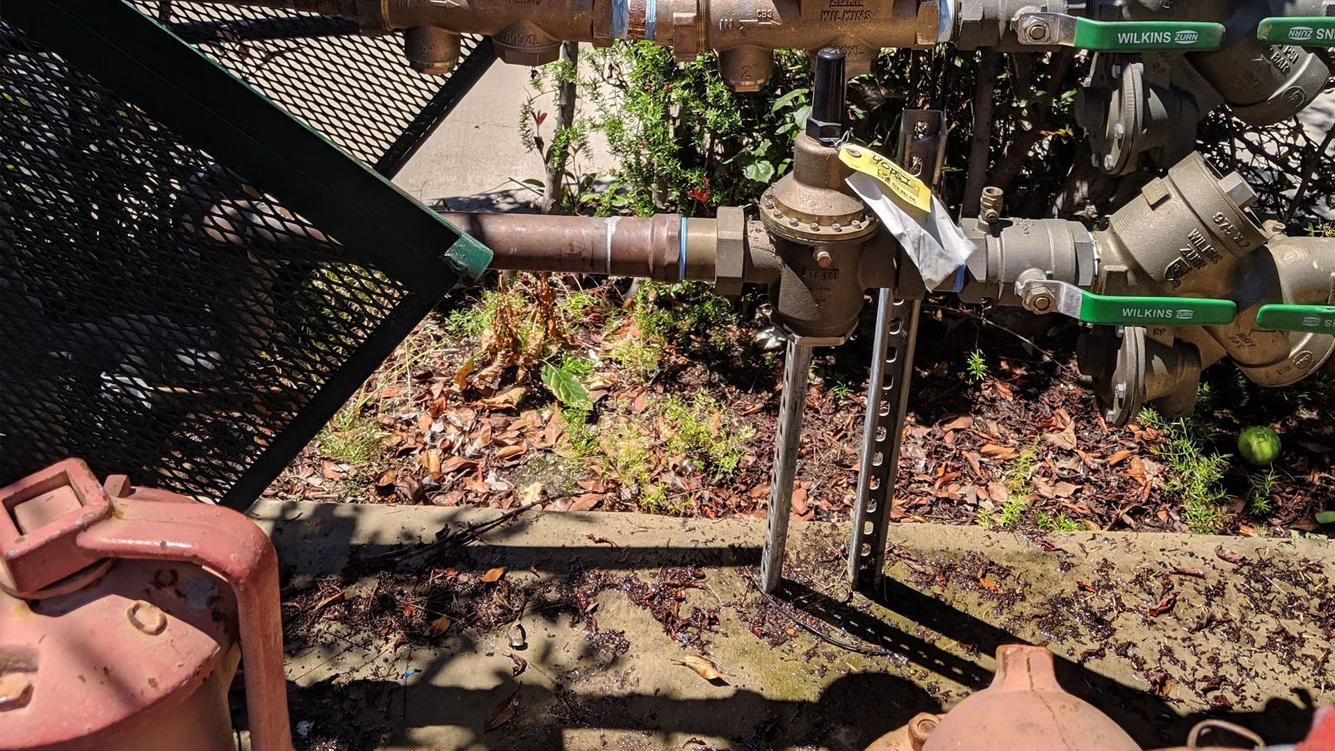 Water pipes and valves outdoors, with a pressure regulator and a small metal support.