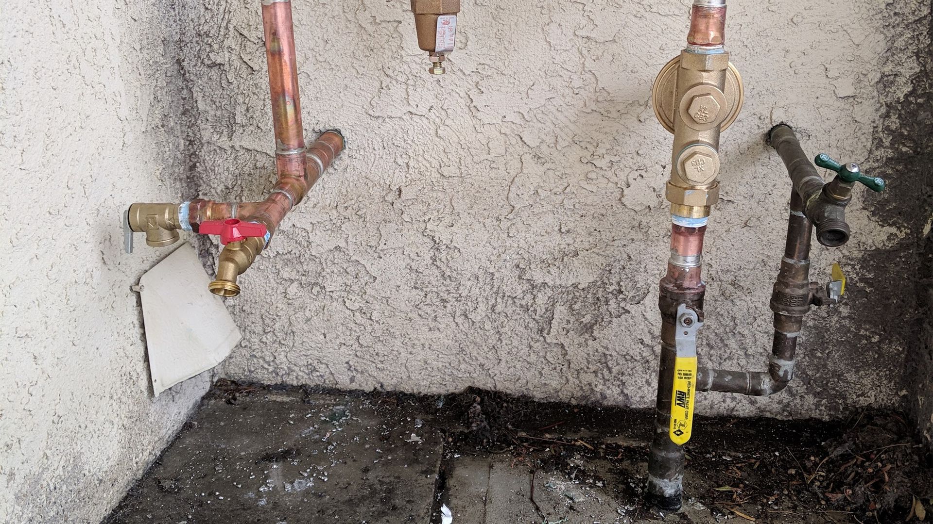 Copper pipes and valves against a textured wall, including a spigot and backflow preventer.
