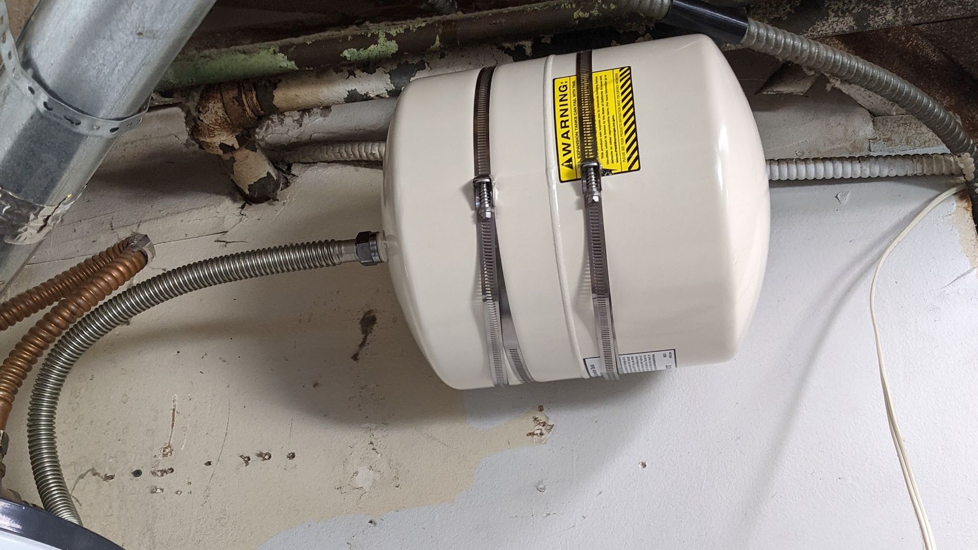 Small beige water heater mounted on a wall with silver hoses attached.