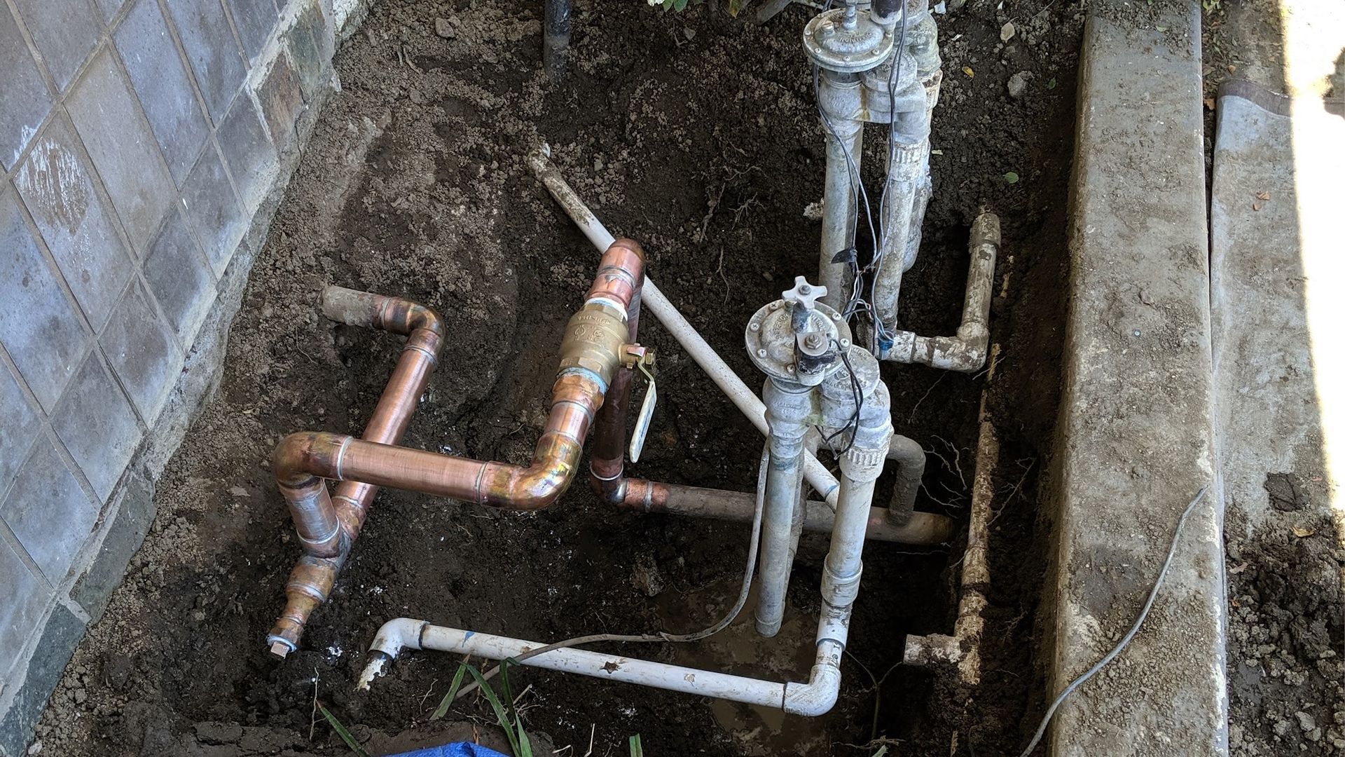Exposed irrigation system with copper and PVC pipes and valves in a dirt-filled trench.