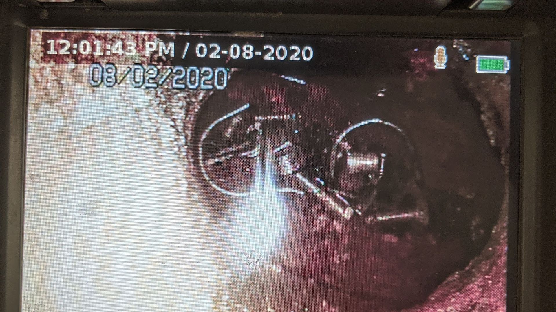 Camera view inside a dark pipe showing metal equipment, wires, and brown liquid. The date 08/02/2020 