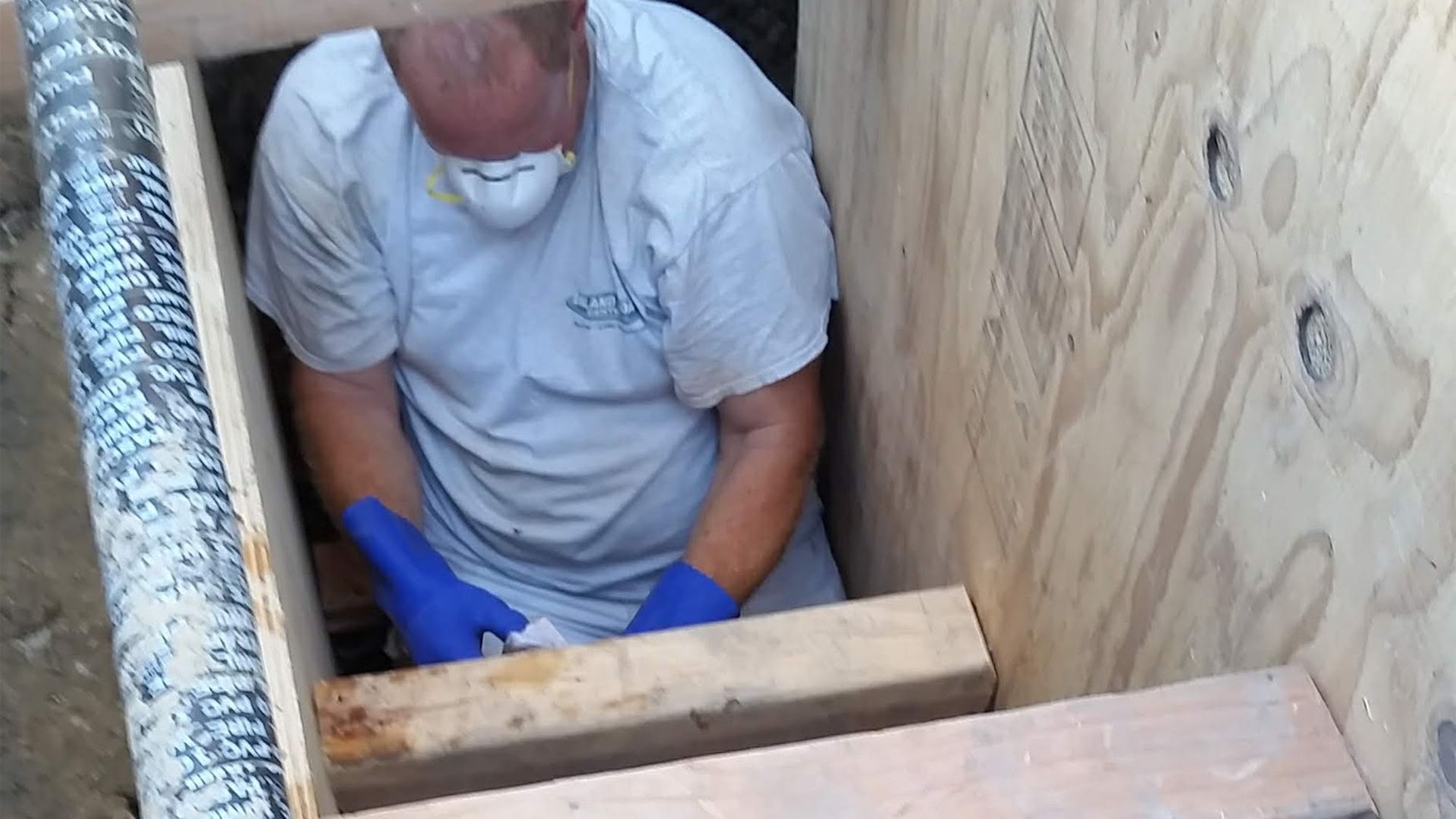 Man in a wooden box, wearing a mask and gloves, working on something unseen.