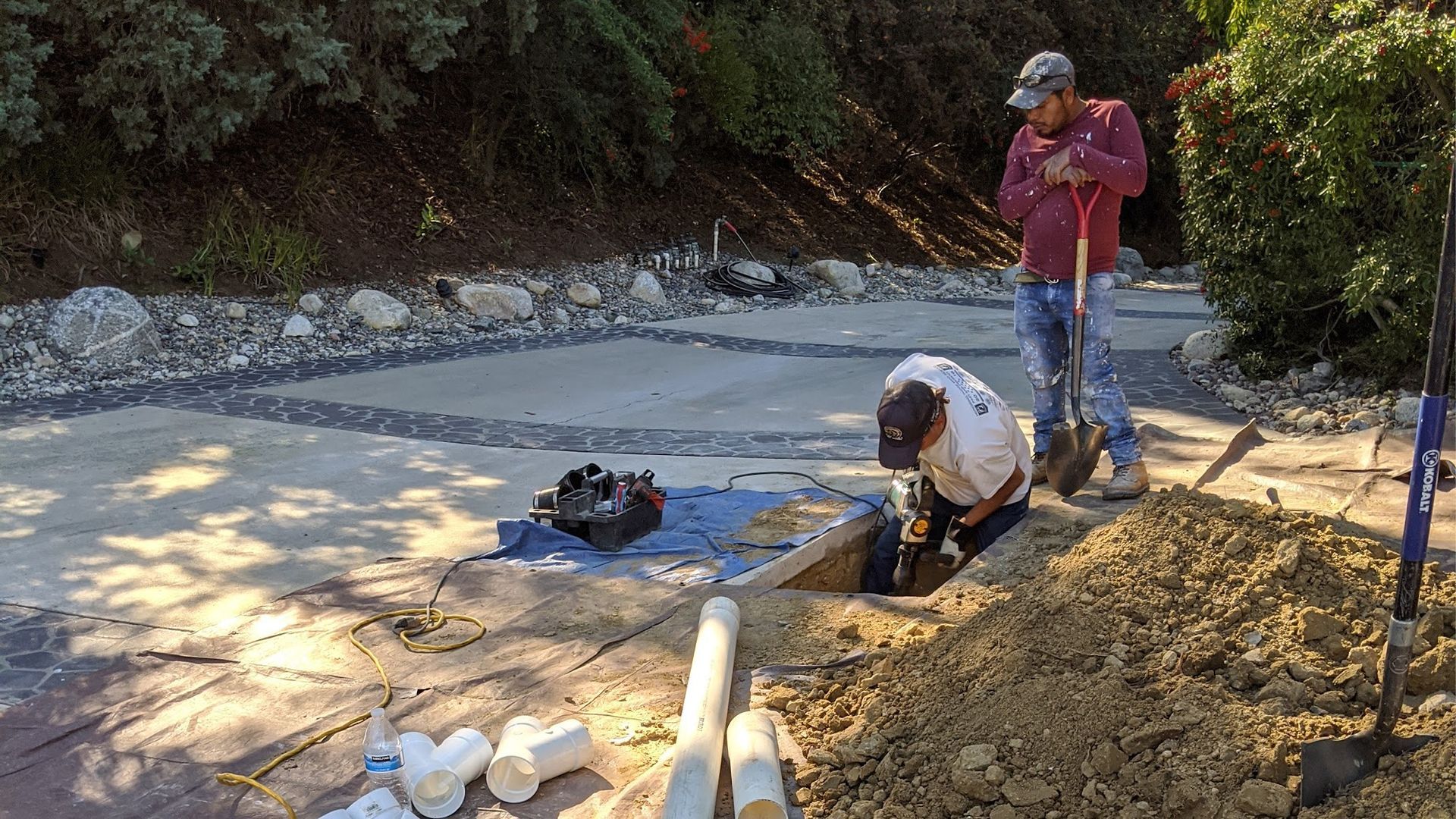 Two workers digging in a paved area, with pipes and equipment nearby. Sunny outdoor setting.