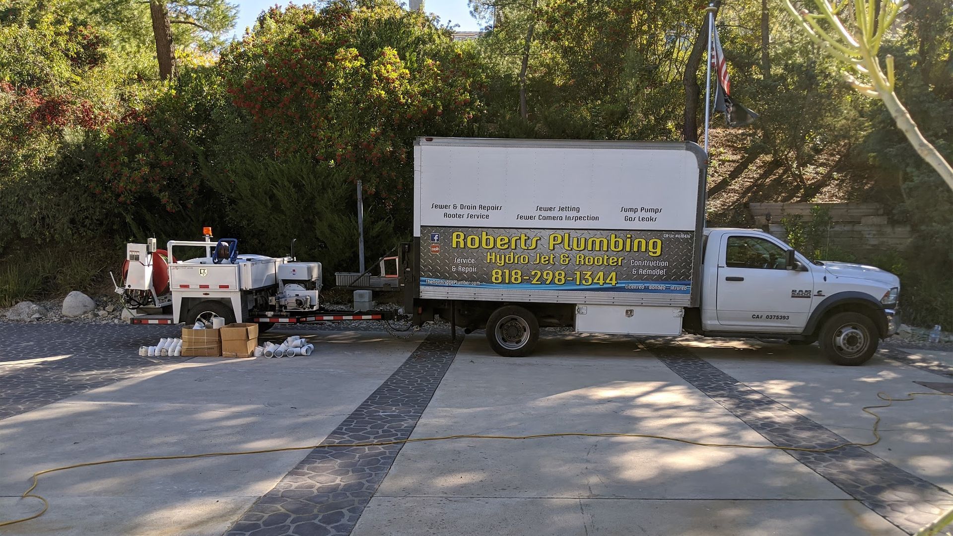 White plumbing truck with attached trailer parked on a concrete surface, near foliage and a stone wall.