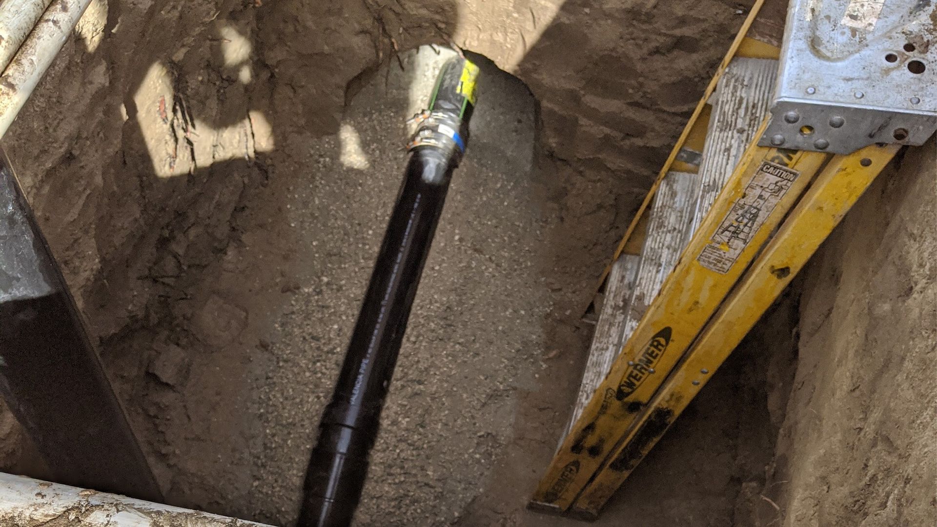 Black pipe in a trench, next to a yellow ladder. Soil visible in the open excavation.