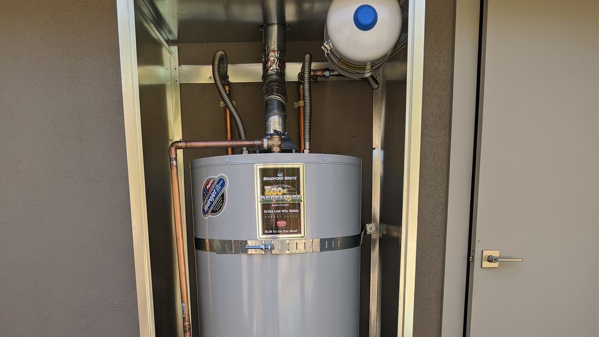 A gray water heater within a metallic enclosure next to a closed beige door.