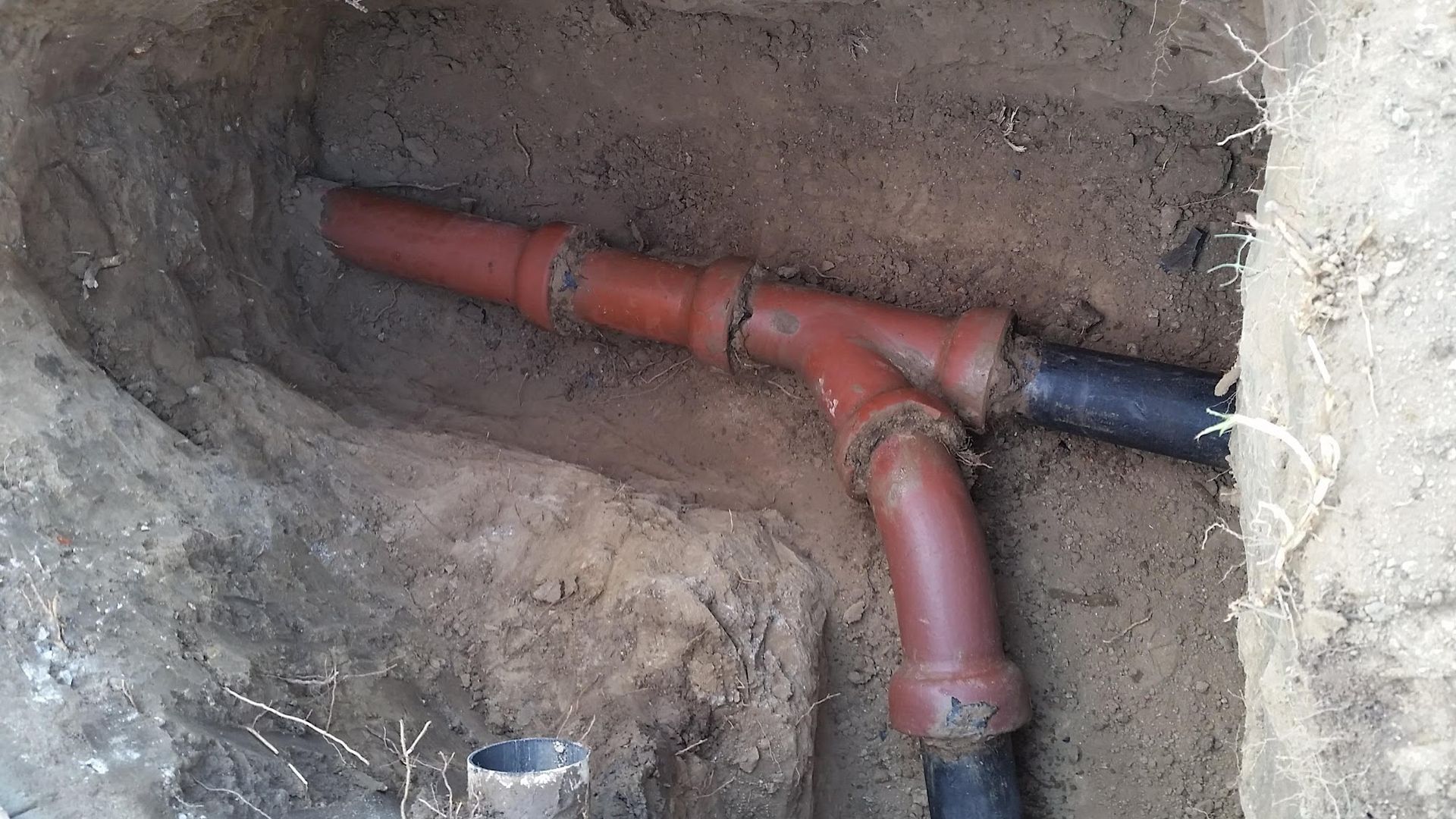 Underground pipes in a trench, connected with reddish-brown and black fittings, surrounded by dirt.