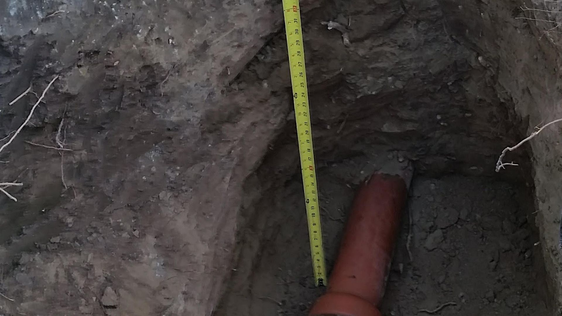 A trench in the ground with an orange pipe. A yellow measuring tape is in the trench.