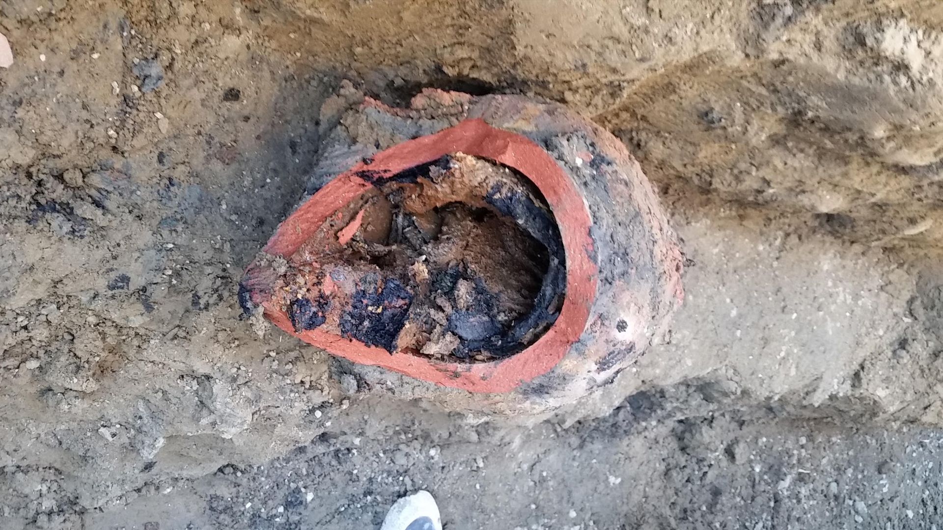 Broken earthenware pipe in sandy soil, red rim, dark interior filled with debris.