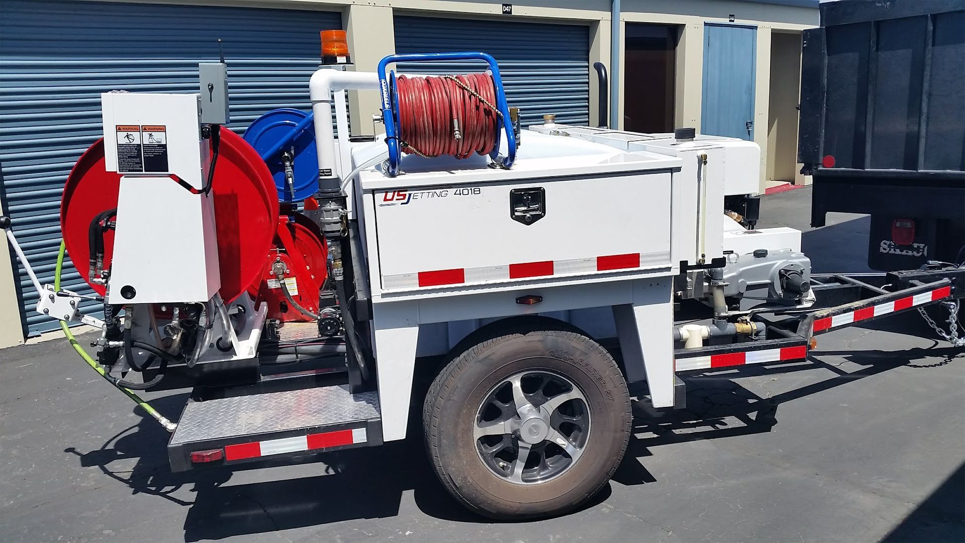 White utility trailer with red/blue reels, a hose reel, and lights in front of a building.