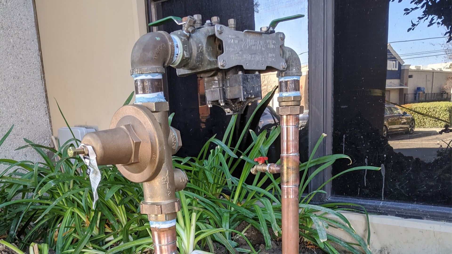 Brown and copper water backflow preventer with green plants in front of a building.