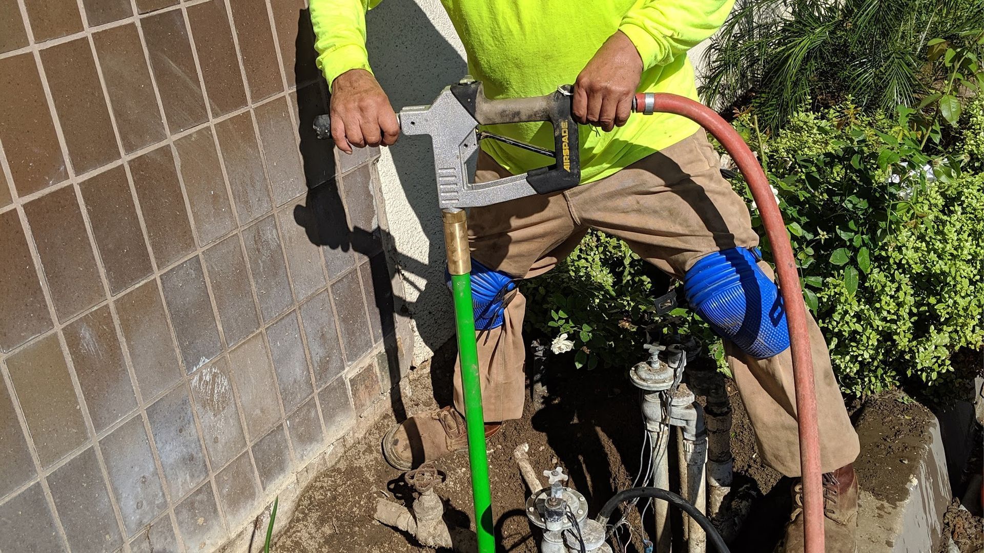 Person in safety gear using a concrete saw near a tiled wall and garden.