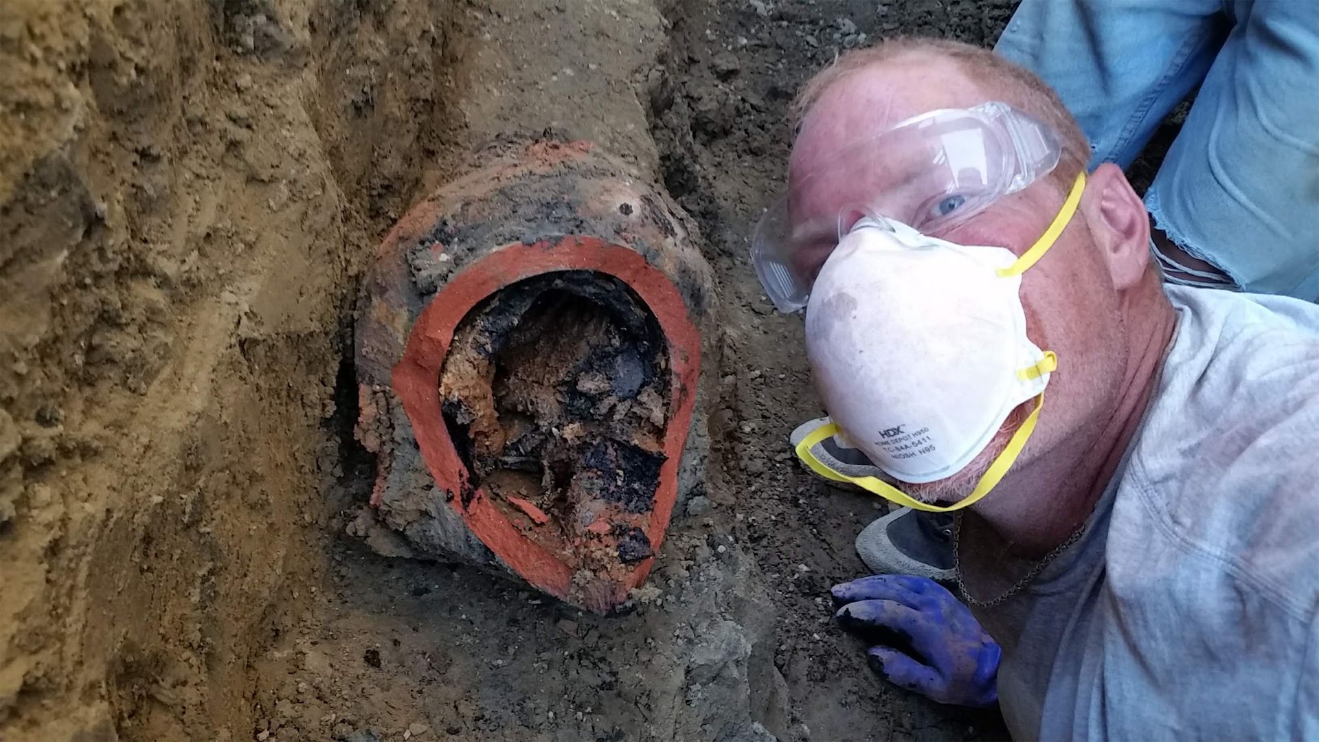 Man in protective gear inspecting a cut, old pipe in a trench. Brown, orange, and gray tones.