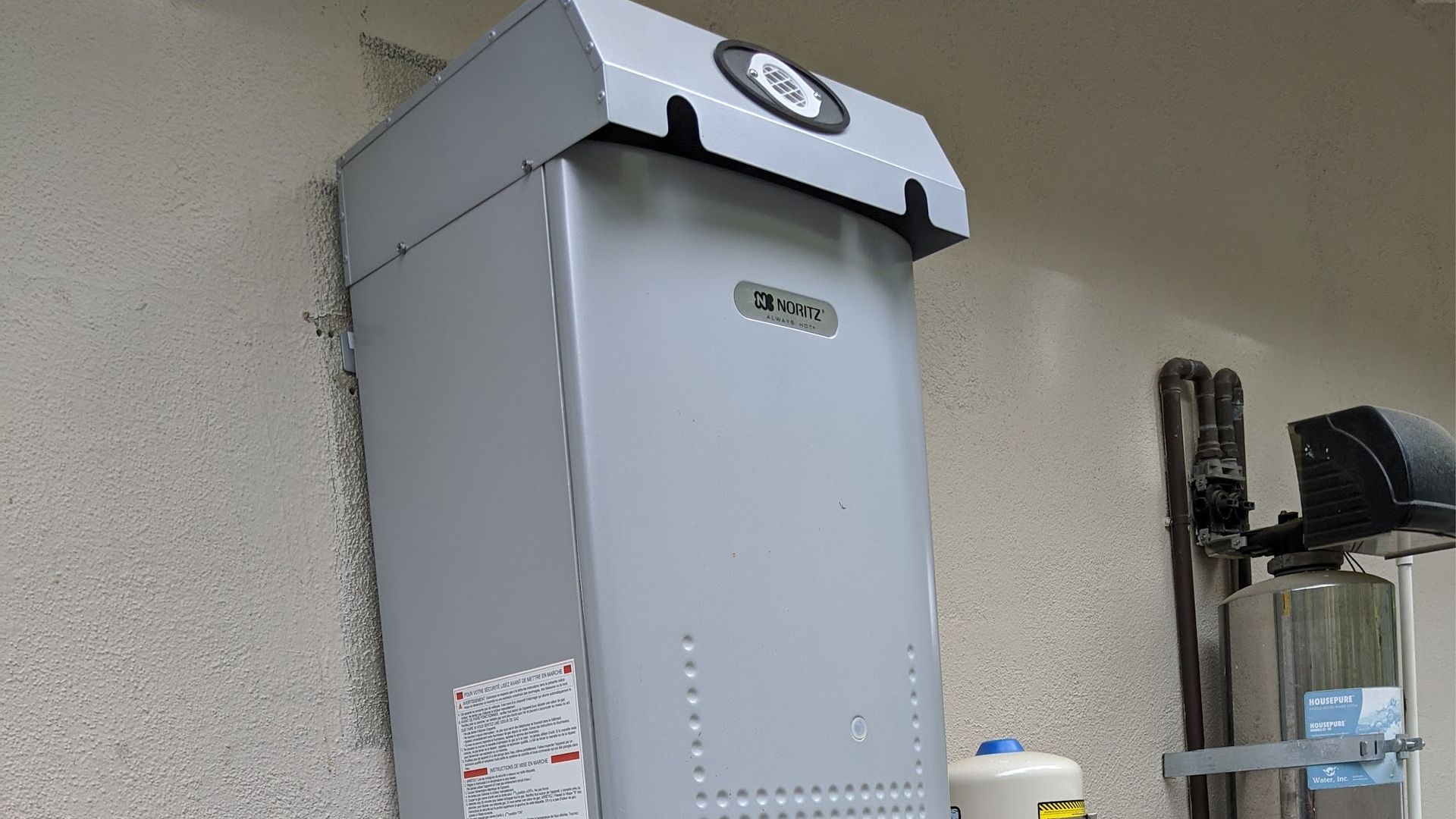 A gray water heater mounted on a stucco wall. A water softener is to the right.