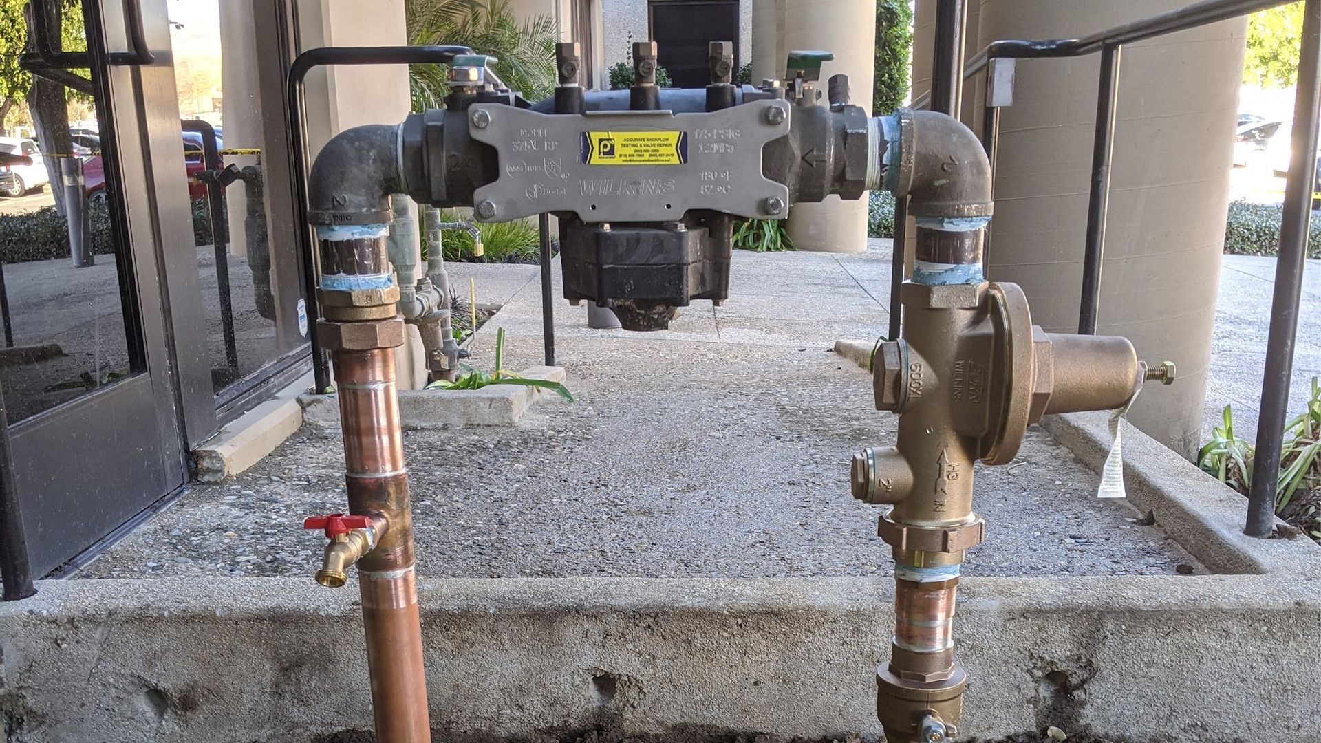 Water system components: pipes, valves, and a pressure regulator, on a gravel surface.