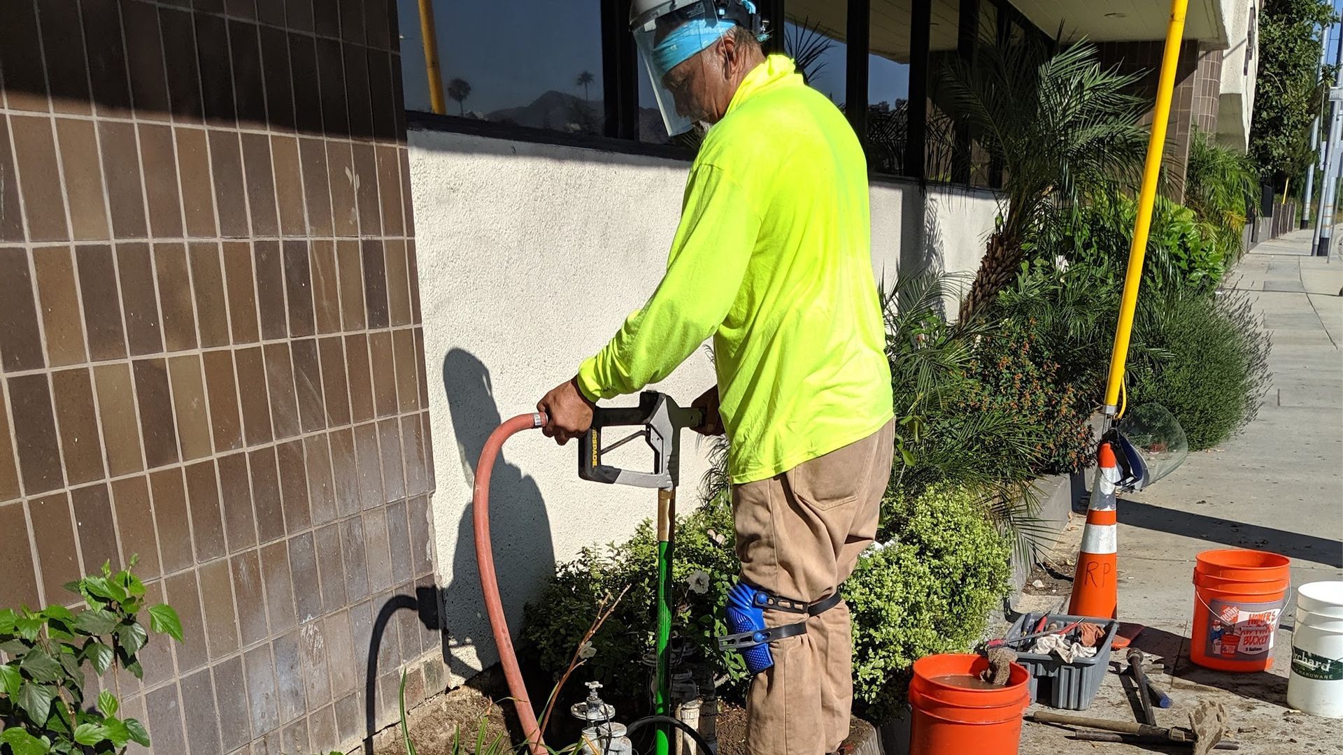 Worker in neon yellow shirt uses a tool near a building. Orange buckets and a pole are nearby.