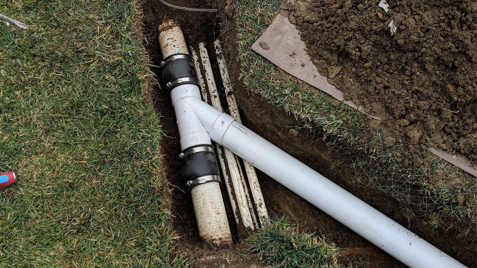 Trench with white and brown pipes joined by black clamps, located on grass next to dirt.