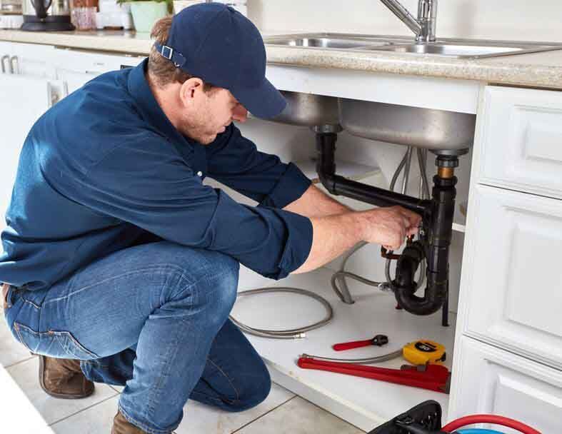 Plumber kneels under a kitchen sink, working on the pipes.