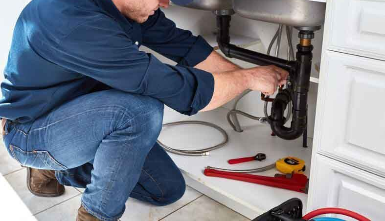A person in a blue shirt and jeans kneels under a kitchen sink, repairing the plumbing with tools laid on the floor.