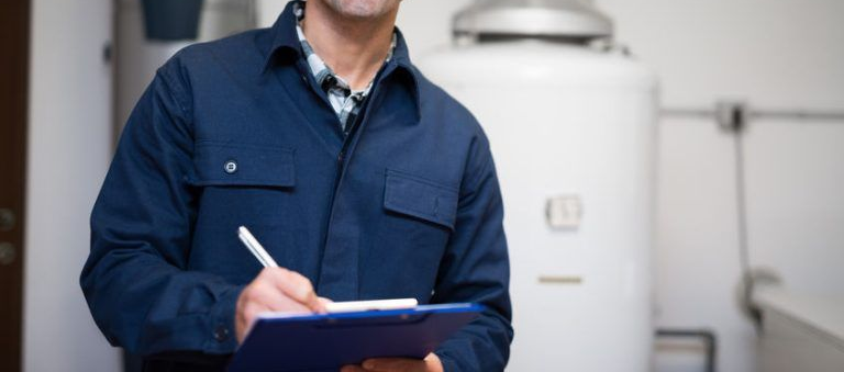 Smiling technician in blue uniform, cap, and clipboard near a water heater and pipes.