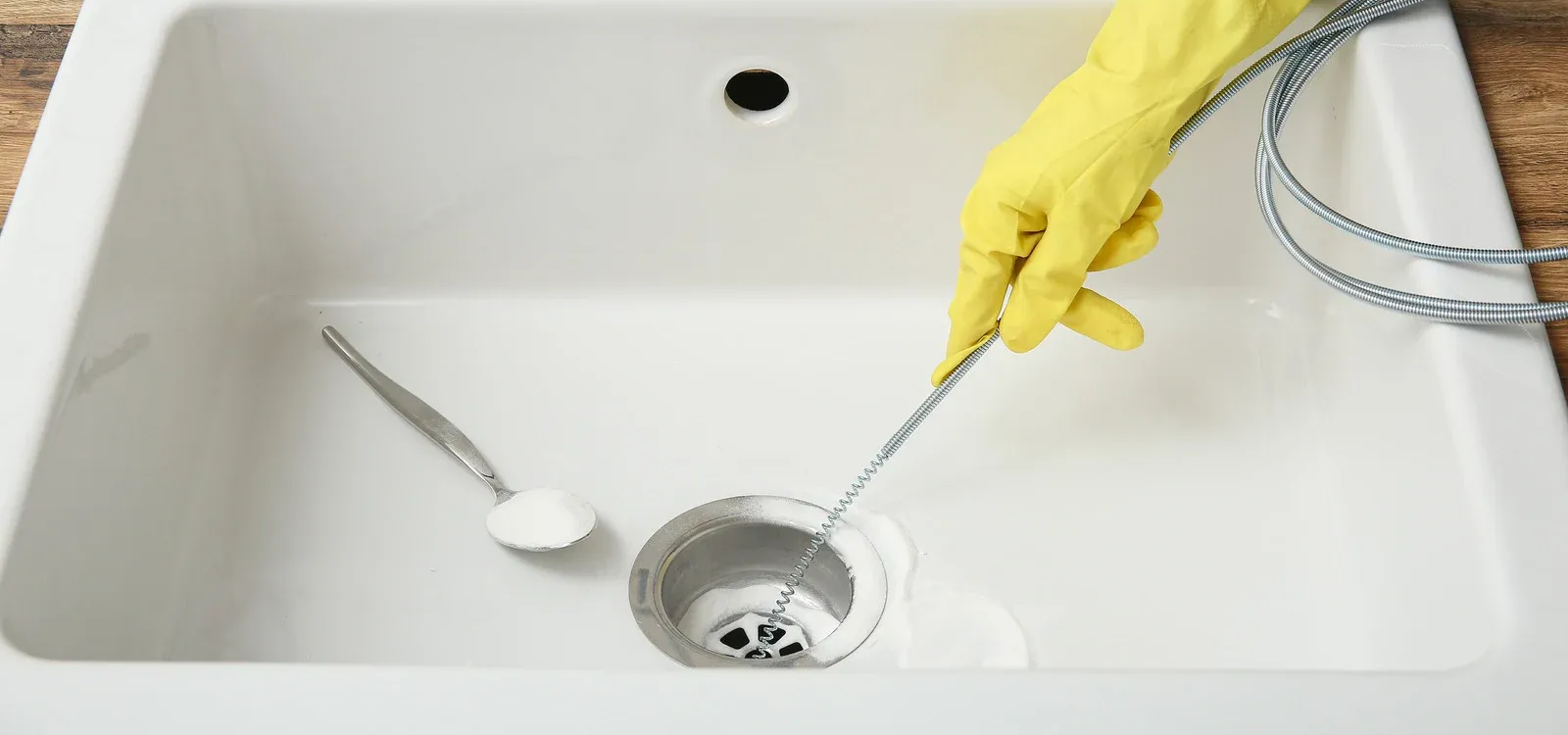 Person in yellow gloves uses a drain snake to unclog a white sink. A spoon with white powder rests nearby.
