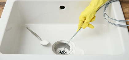 Person in yellow gloves uses a drain snake to unclog a white sink. A spoon with white powder rests nearby.