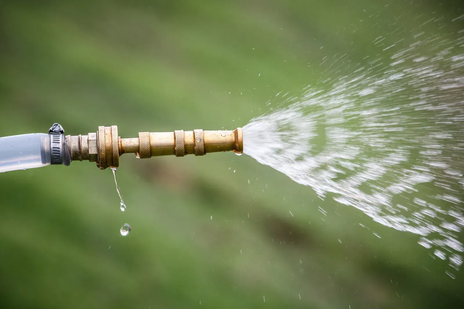 Water spraying from a brass hose nozzle, drops falling, against a blurred green background.