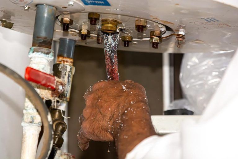 Hand inspecting a leaking water heater element. Water spraying, white coating present.