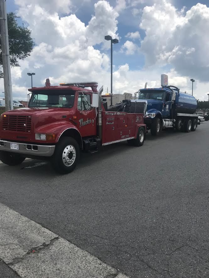 Red tow truck pulling a blue tanker truck on a paved lot under a cloudy sky.