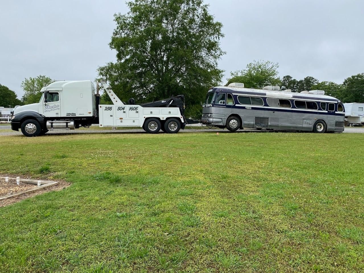Tow truck pulling a silver and blue bus on a grassy field, overcast sky.
