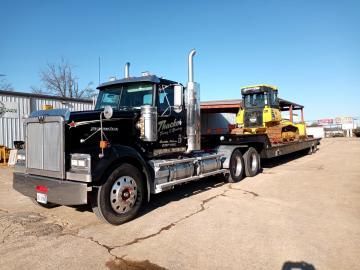 Black semi-truck hauling a yellow construction vehicle on a flatbed trailer on a sunny day.
