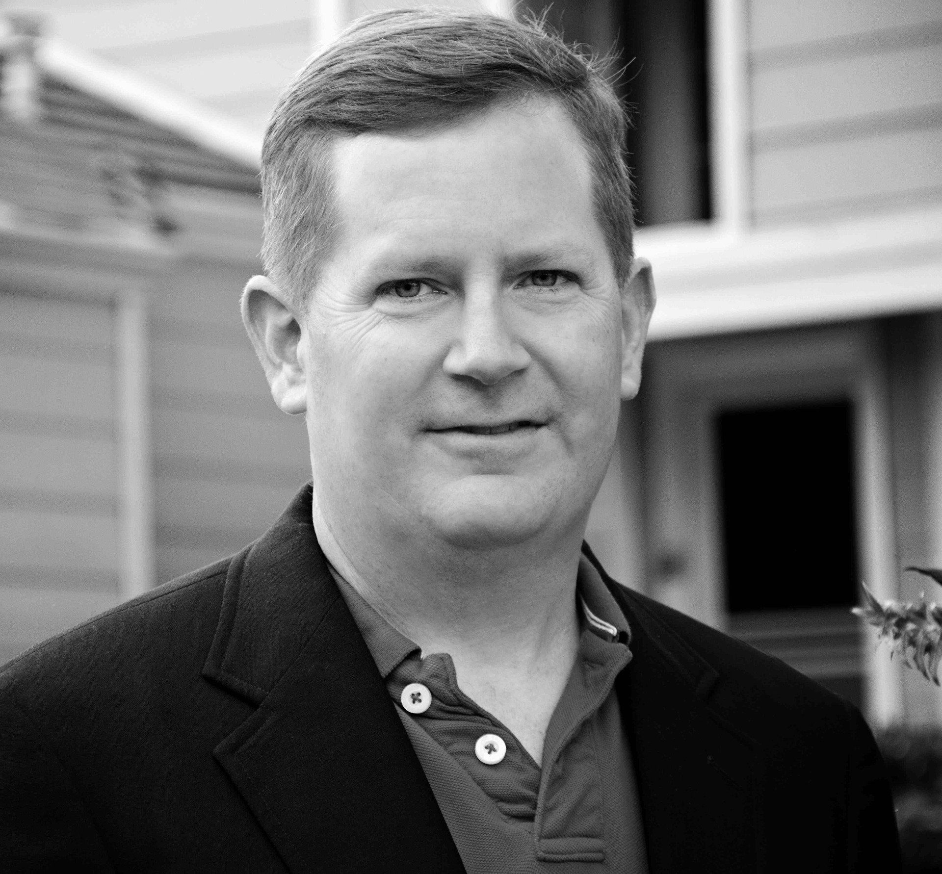 A man in a suit is standing in front of a house in a black and white photo.