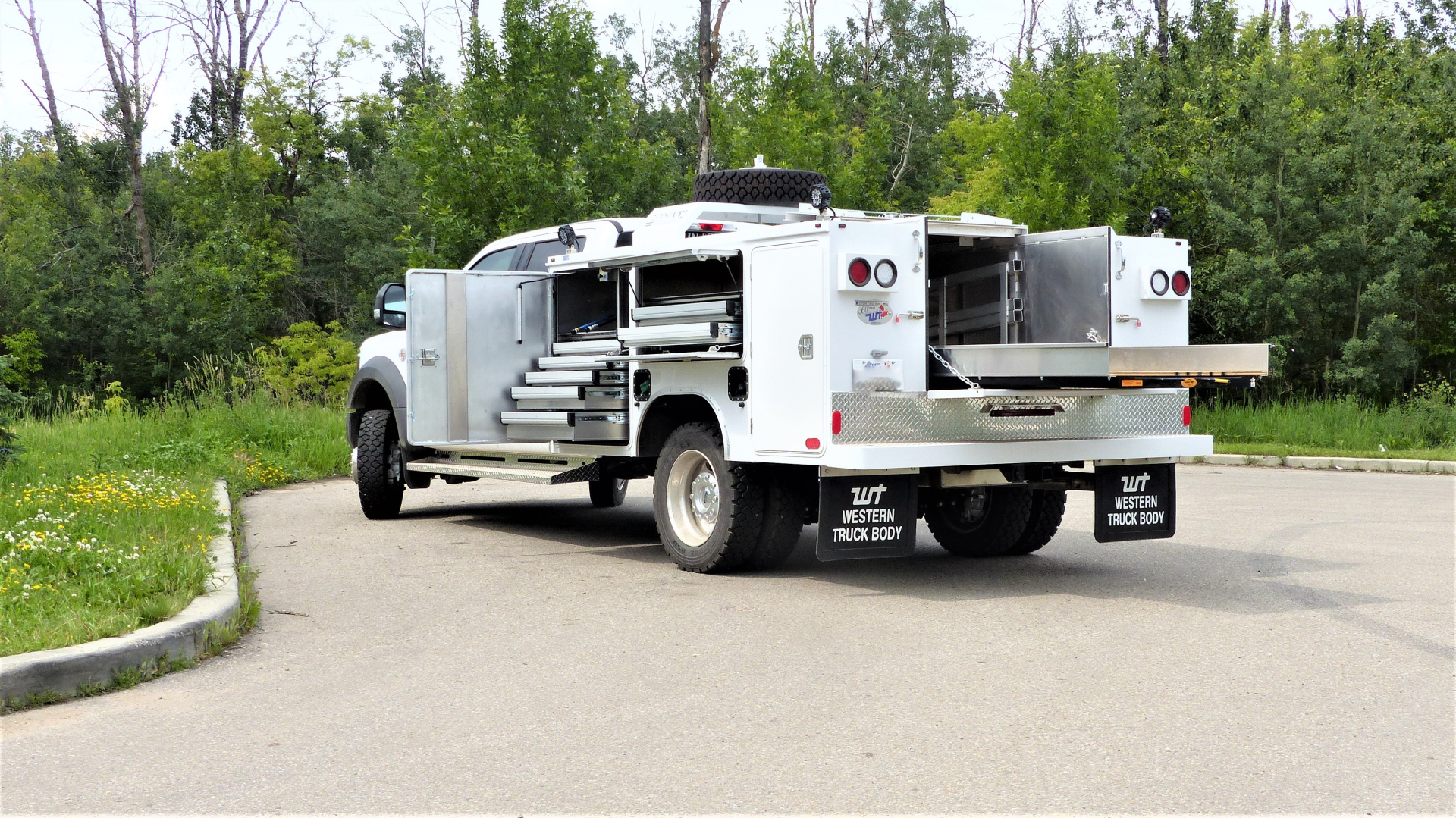 A white service truck with open storage compartments and extended tool drawers, parked on a paved lot near trees.