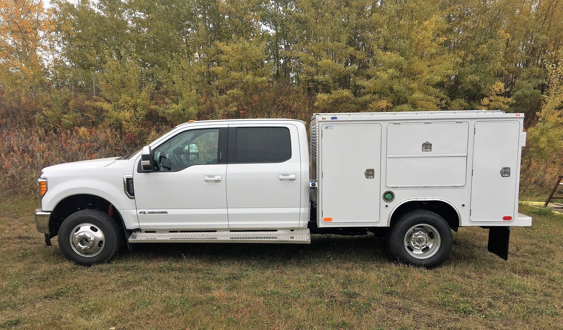 A white crew-cab utility truck parked on a grass field with autumn trees in the background.