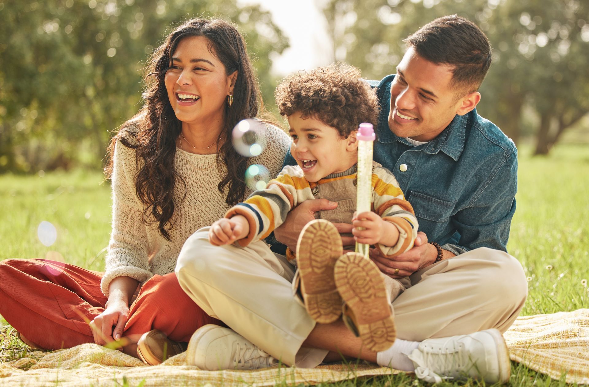 Family picnicking in a park; child laughs with bubbles, parents smile. Sunny day on a blanket.