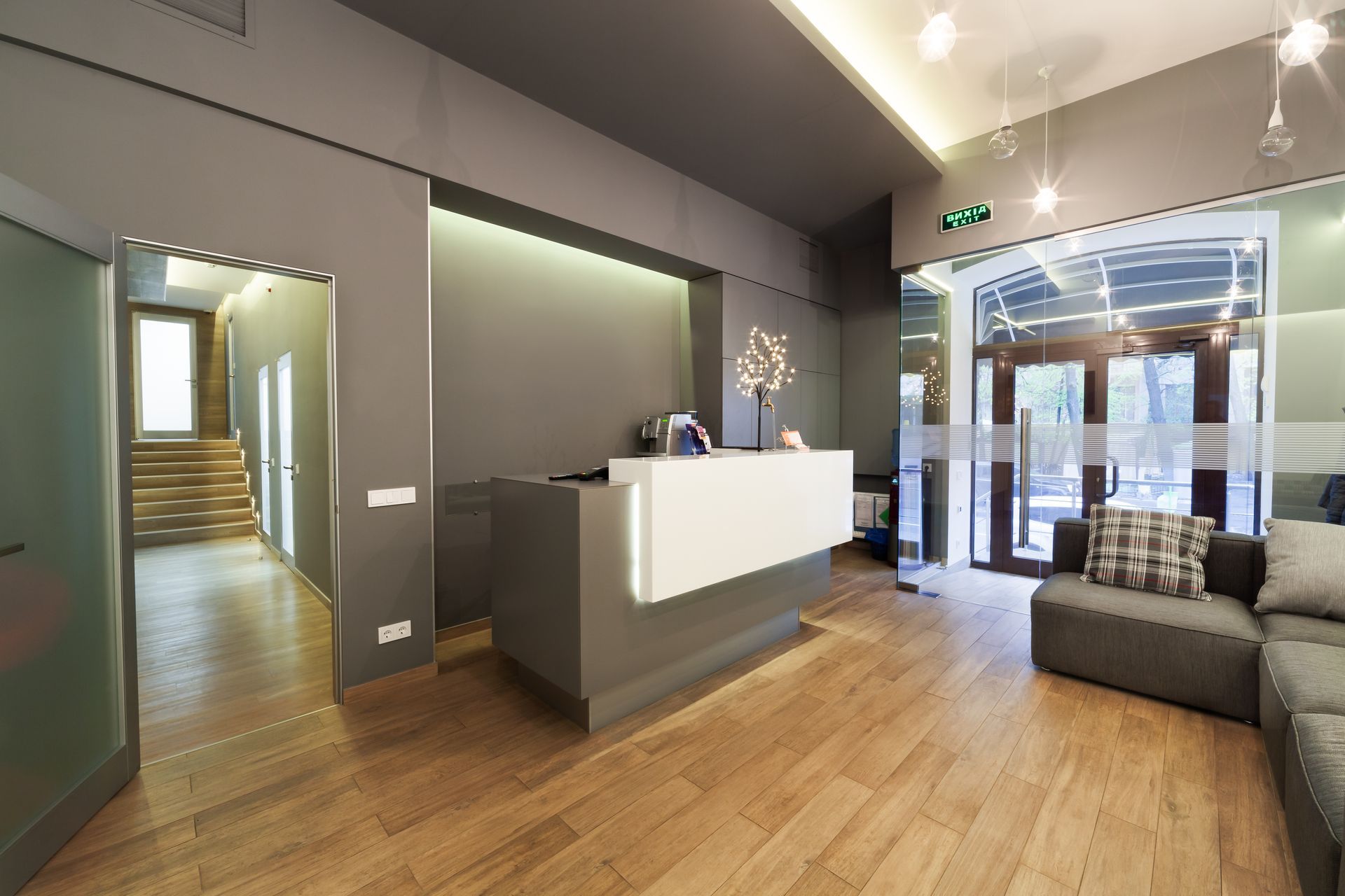 Modern hotel lobby with gray walls, wood floors, white reception desk, and glass entrance.