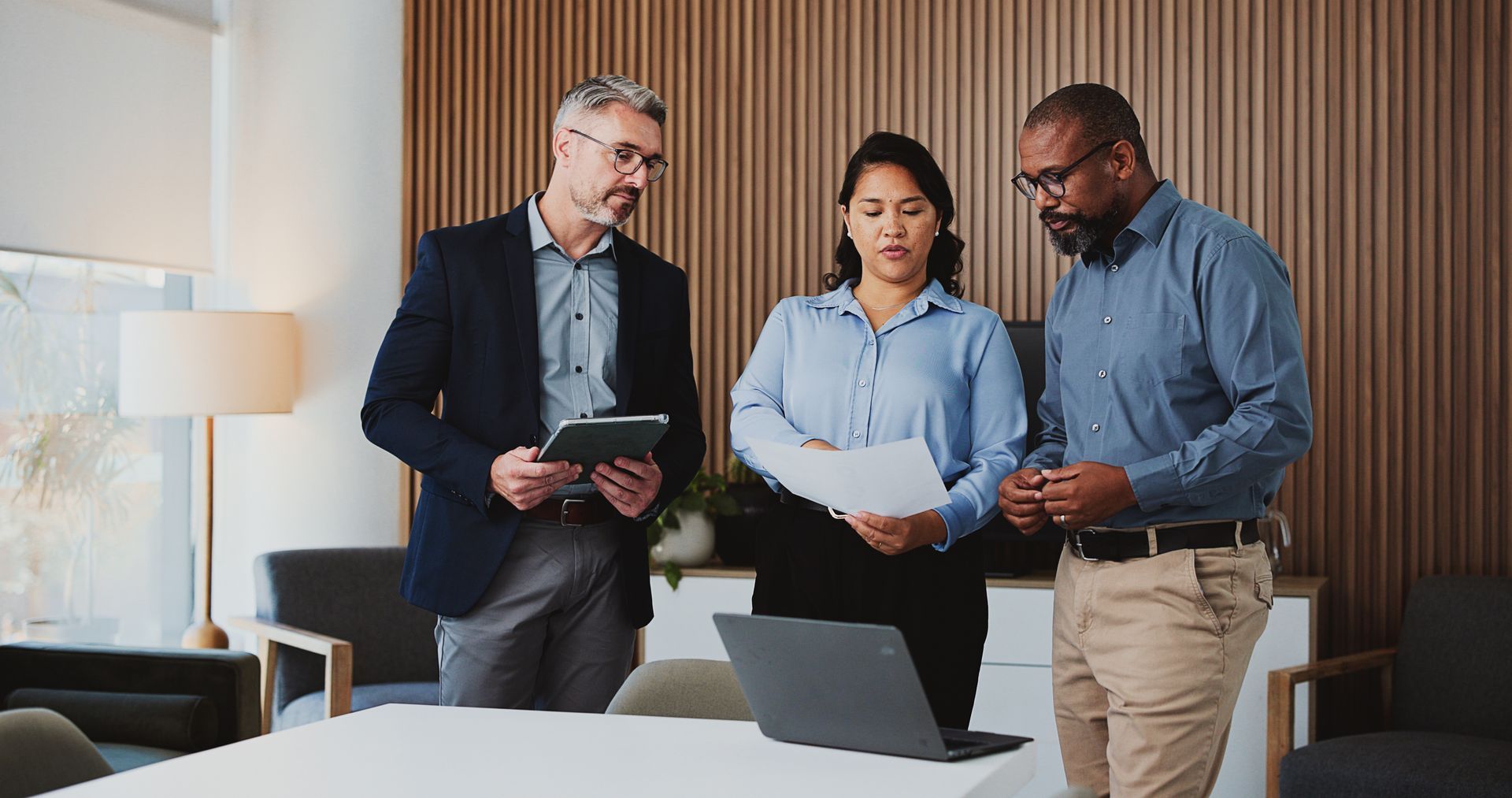 Three coworkers reviewing documents around a laptop in a modern office.