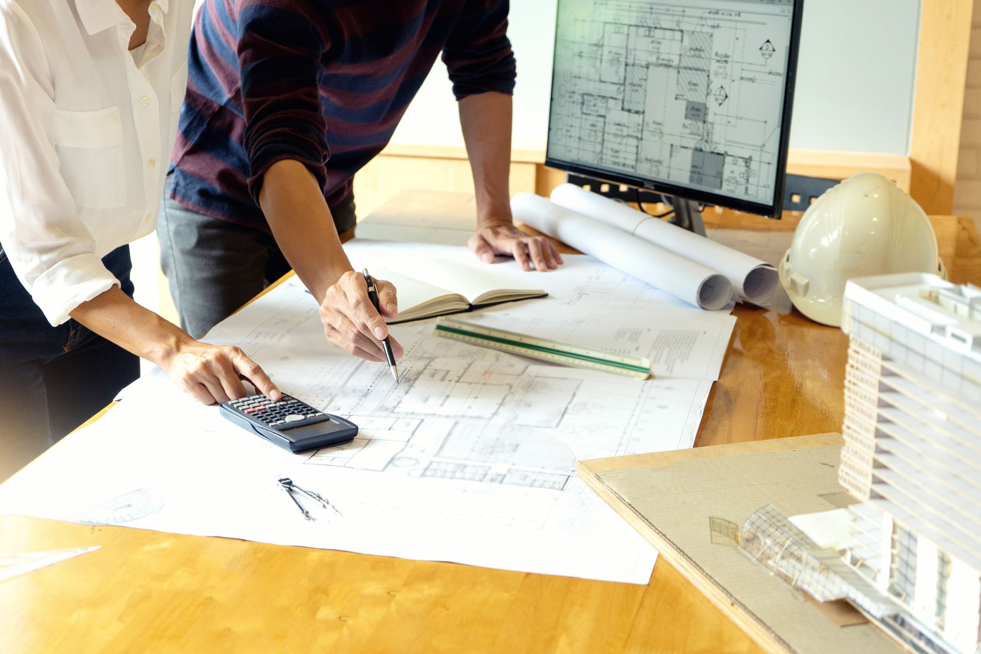 Two people reviewing blueprints at a desk with a calculator, ruler, and computer monitor.